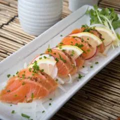 Tataki de salmón fresco, finamente cortado y adornado con rodajas de limón, rábano y cebollino, servido sobre una cama de finas hebras de cebolla.