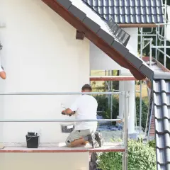 Dwóch malarzy na rusztowaniu odnawia elewację domu. Używają wałków i wiadra z farbą malarską rozpuszczalną w wodzie.