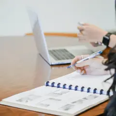 Ręka pisze notatki, przygotowując się do napisania podania o przedłużenie umowy o pracę. Obok laptop i telefon.
