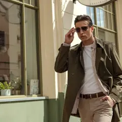 Mężczyzna w eleganckim, khaki płaszczu i beżowych spodniach poprawia okulary, idąc ulicą.
