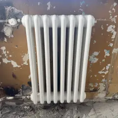 Biały grzejnik żeberkowy zamontowany na zniszczonej, brązowej ścianie.