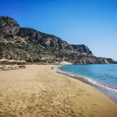 Piękna, piaszczysta plaża Tsambika z turkusową wodą i skalistym wzgórzem w tle. Leżaki i parasole czekają na plażowiczów.
