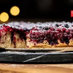 Pyszne ciasto z porzeczkami, posypane cukrem pudrem, na ciemnym talerzu. W tle rozmyte, ciepłe światła.