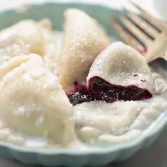 Pyszne pierogi z jagodami, polane śmietaną i posypane cukrem, czekają na widelca.