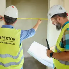 Dwóch inspektorów w kaskach i kamizelkach mierzy ścianę taśmą. To symboliczne pierwsze klucze do nowego domu.