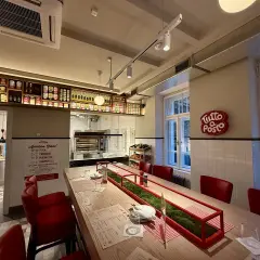 Wnętrze restauracji z czerwonymi krzesłami i długim stołem. Na ścianie wisi neon "Tutto Posto", a nad ladą półki z produktami. Otto Pompieri opinie: warto odwiedzić!