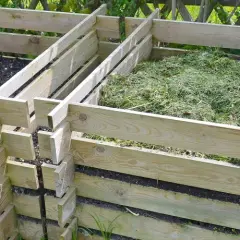Holzkomposter mit zwei Fächern, eines gefüllt mit Erde, das andere mit frisch gemähtem Gras, bereit zum Kompost zerkleinern.