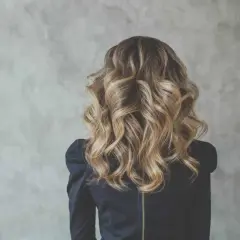 Kobieta z burzą falowanych, **bronde włosów do ramion**. Złociste refleksy pięknie mienią się na tle szarej ściany.