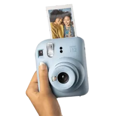 Niebieski aparat Instax Mini 12, trzymany w dłoni, z właśnie wysuniętym, jasnym zdjęciem dwóch uśmiechniętych przyjaciółek.