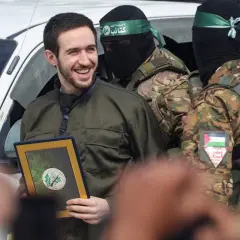 Uśmiechnięty mężczyzna w zielonej kurtce odbiera ramkę z dokumentem, otoczony przez uzbrojonych bojowników. To symboliczna chwila, być może związana z wyzwoleniem (liberation po polsku).