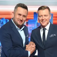 Dwóch mężczyzn w garniturach ściska sobie dłonie na tle scenografii z miejskim krajobrazem, gdzie nagrywane jest koło fortuny.