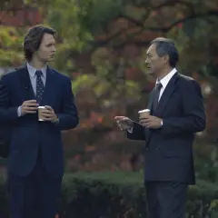 Zwei Männer in Anzügen, die Kaffee halten, diskutieren im Freien. Ein Mann ist jünger, der andere älter. Szene aus Tokyo Vice Staffel 2.