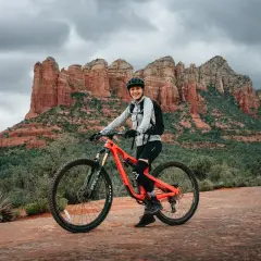 A smiling woman in riding gear poses on her bright orange mountain bike, ready for a trail tune-up amidst the stunning red rock formations of Sedona.