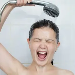 Une femme crie sous une douche froide, visiblement contrariée par un problème d'eau chaude.