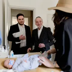 Un bébé est allongé sur une table, une tétine bleue dans la bouche. Des hommes en costume, dont un portant une kippa, observent le déroulement de la circoncision juive.
