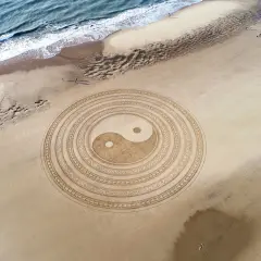 Símbolo Yin Yang en la arena, representando la energía masculina y femenina en equilibrio. El mar y la playa completan la escena.