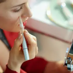 Kobieta aplikuje korektor pod oczy za pomocą pędzla, patrząc w lusterko.