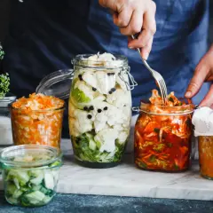 Här är burkar med fermenterad mat: blomkål, morot och kål. En hand använder en gaffel för att plocka från en burk.