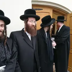 Chasydzi w tradycyjnych strojach, czarnych kapeluszach i z pejsami, stoją przed drewnianymi drzwiami.