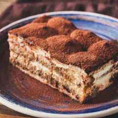 Une part généreuse de tiramisu cake, saupoudrée de cacao, repose sur une assiette bleue. Un délice italien.