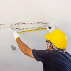 Un homme en casque jaune mesure des taches d'humidité au plafond, cherchant une solution humidité maison.