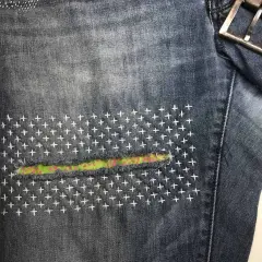 Close-up of denim jeans with a colorful fabric patch and white cross-stitch embroidery, showcasing a stylish sashiko jeans repair.