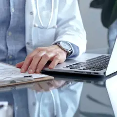 Lekarz w białym kitlu pisze na dokumentach, obok laptop. Szybka konsultacja psychiatryczna online.