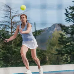 Kobieta w białej spódniczce i niebieskiej koszulce ćwiczy tenis przy ścianie, odbijając piłkę rakietą.