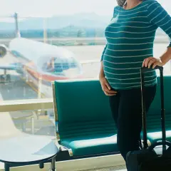 Czekając na lotnisku, przyszła mama zastanawia się, czy w ciąży można latać samolotem. Obok walizka, za oknem samolot.