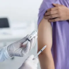 Rękawiczki medyczne trzymają fiolkę z preparatem i strzykawkę. Osoba w fioletowej koszulce przygotowuje się do szczepienia, być może przeciwko duru brzusznemu.