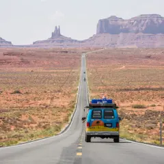 Błękitny van z bagażem na dachu jedzie pustynną drogą w USA. Monument Valley w tle. To część ich wycieczki do USA.