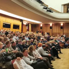 Widownia w teatrze, ludzie siedzą w rzędach, ubrani elegancko.
