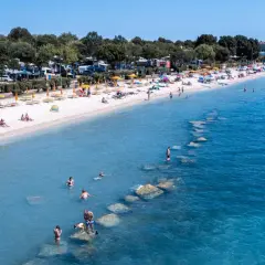 Słoneczna plaża z turystami, parasolami i kamperami w tle. Woda kusi do kąpieli, a kamienie tworzą naturalne falochrony. Idealne miejsce na bi village camping.
