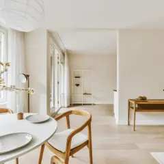 Helles Esszimmer mit weißem Tisch, Holzsessel und Blumen. Ein Regal und ein Beistelltisch mit Deko unterstreichen den "weniger ist mehr lebensstil".