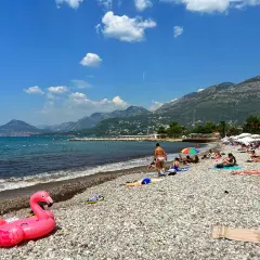 Słoneczny dzień na plaży w Barze, Czarnogóra. Ludzie opalają się, a w tle widać góry. Różowy flaming unosi się na kamienistej plaży.