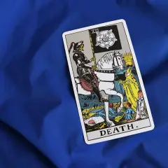 Tarot karta Śmierci na niebieskim tle. Postać w zbroi na białym koniu, niosąca flagę z różą, symbolizuje nieuchronne zmiany i transformację.