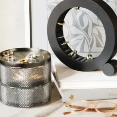 Un joyero vertical circular negro exhibe anillos dorados. A su lado, un joyero cilíndrico plateado con dos niveles.
