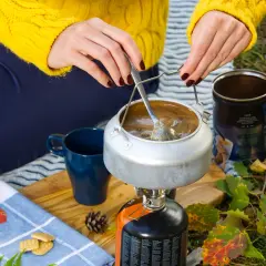 Preparando café en la naturaleza con recetas camping gas. Una tetera hierve sobre un hornillo, junto a una taza azul y galletas.