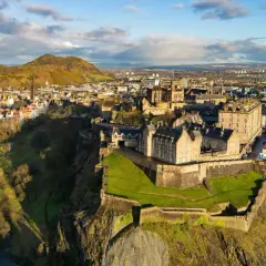 Edinburgh Castle tronar över staden, Skottlands huvudstad. En historisk vy med gröna kullar och moln på himlen.