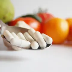 Łyżka z białymi kapsułkami na tle owoców i warzyw.