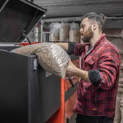 Mężczyzna w flanelowej koszuli wsypuje pellet do pieca. W tle worki z paliwem.