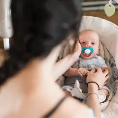 Maluszek w bujaczku polecanym przez fizjoterapeutów, z niebieskim smoczkiem. Mama delikatnie głaszcze go po główce.