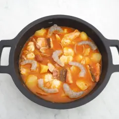 Un delicioso guiso marinero burbujea en una cazuela negra, con patatas, gambas, pescado y almejas en una salsa rojiza.