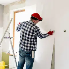 Mann mit roter Kappe und Handschuhen kleistert Vliestapete ein, um eine Wand zu tapezieren.