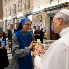 Eine Ordensfrau überreicht dem Papst ein Geschenk. Im Hintergrund stehen weitere Ordensschwestern und Männer in Anzügen.