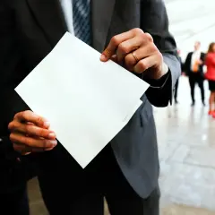 Mężczyzna w garniturze trzyma białą kopertę, jakby miał dokonać przekierowania. W tle ludzie w eleganckich strojach.