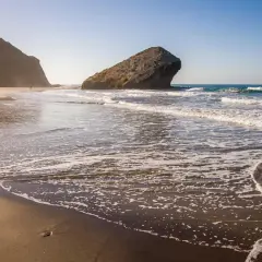 La duna de Monsul se extiende hacia el mar, con olas rompiendo suavemente en la arena dorada. Una figura solitaria camina por la playa.