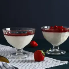 Dwa pucharki z panna cottą, udekorowane musem truskawkowym i owocami leśnymi. Obok leżą świeże truskawki i złota łyżeczka.