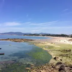 Vista de la playa de A Calzoa en Vigo, con gente disfrutando del sol y el mar. Barcas pequeñas flotan en aguas cristalinas.