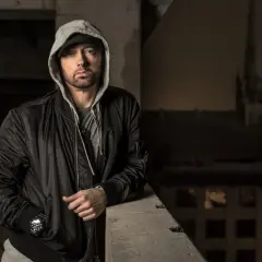 Eminem w czarnej kurtce bomberce i szarej bluzie z kapturem, oparty o betonową barierkę.